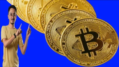 A young man wearing a beige t-shirt, glasses, and a black watch points with both fingers to Bitcoin.