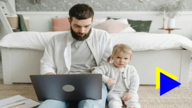 A man sitting in front of the bed wearing a white t-shirt over a gray t-shirt and browsing the laptop with his son next to him