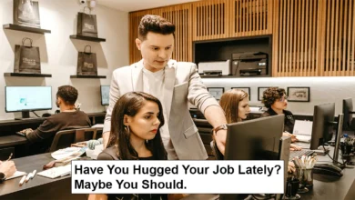 A woman who appears to be an employee is sitting in front of a laptop, and a man who appears to be her manager is directing her. In the background are employees.
